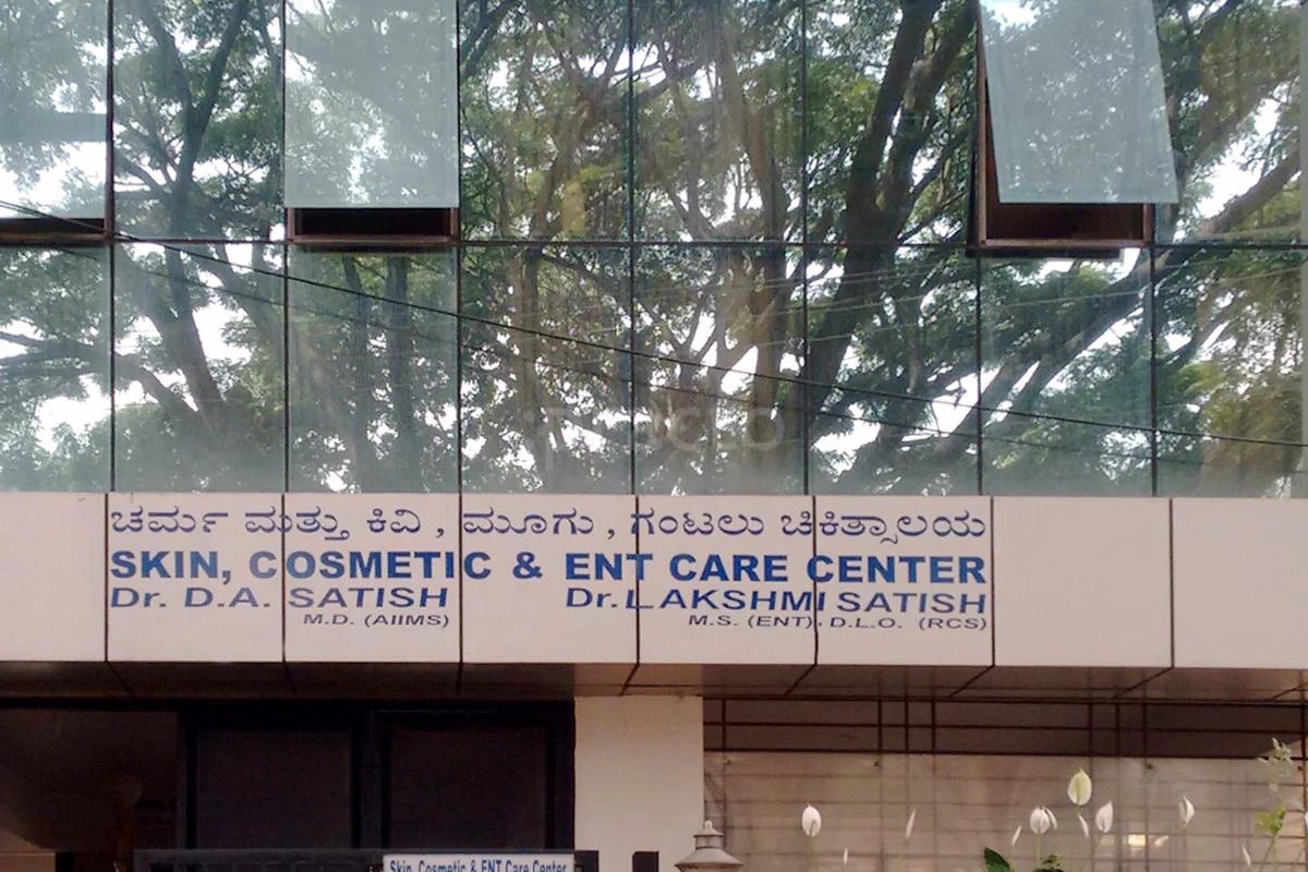 The front of the clinic building, with the signboard in Kannada and English reading Skin, Cosmetic & ENT Care Center, Dr. D. A. Satish and Dr. Lakshmi Satish, below a glass facade reflecting the trees.