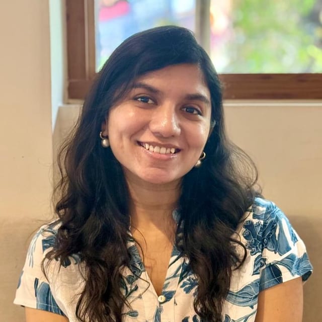 Photograph of Dr. Amrutha Herle P M seated on a beige sofa beside a window, wearing a white and blue printed dress.