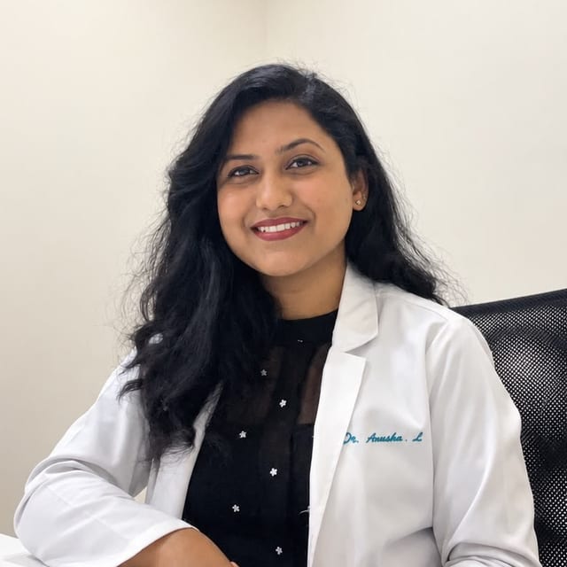 Photograph of Dr. Anusha L seated at a white desk in a consulting room, wearing a white coat embroidered with her name over a black top, hands resting on the desk.