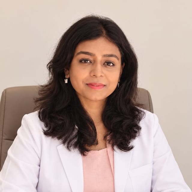 Photograph of Dr. Tushara Jaiprakash seated at a desk in an office chair, wearing a white coat over a pink top, hands folded on the desk, against a pale grey wall.